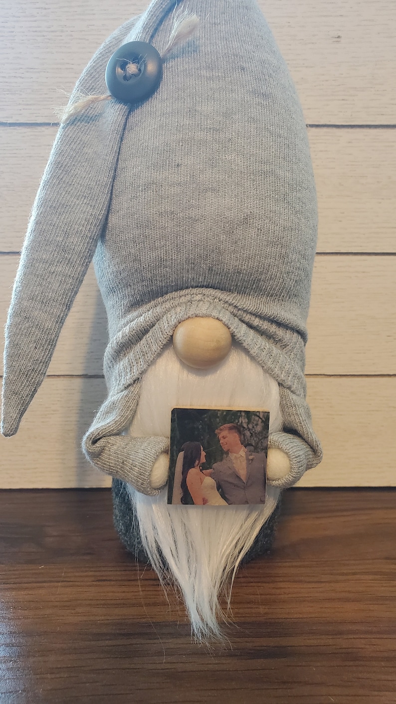 Puede incluir: Un gnomo gris con barba blanca sosteniendo una peque&ntilde;a foto de una novia y un novio. El gnomo lleva un gorro gris con un bot&oacute;n negro.