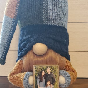 Puede incluir: Un gnomo con un gorro de punto azul y marr&oacute;n, barba marr&oacute;n y nariz de madera sostiene una foto enmarcada de una familia. La foto muestra a una mujer, un hombre y una ni&ntilde;a.
