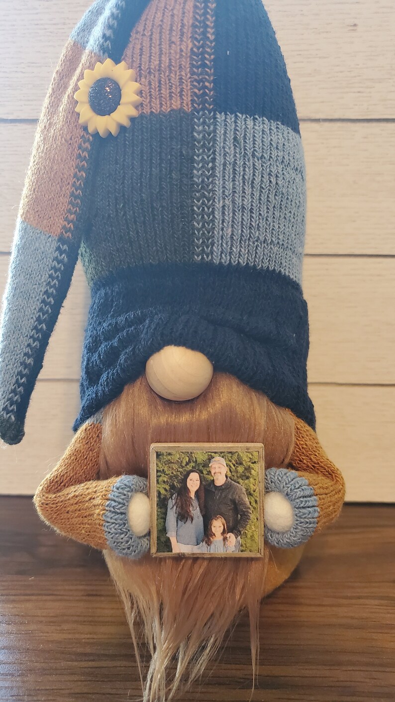 Puede incluir: Un gnomo con un gorro de punto azul y marr&oacute;n, barba marr&oacute;n y nariz de madera sostiene una foto enmarcada de una familia. La foto muestra a una mujer, un hombre y una ni&ntilde;a.