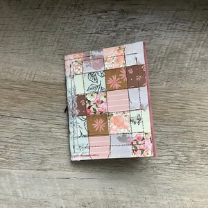 May include: A small, pink and brown fabric-covered notebook with a patchwork design featuring floral and musical note patterns.