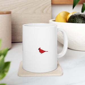 May include: White ceramic coffee mug with a red cardinal bird illustration. The mug has a handle and is sitting on a beige coaster.