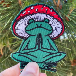 May include: Green frog embroidered patch with closed eyes, sitting in a meditative pose with hands clasped in front of its chest. The frog is wearing a red and white mushroom cap.