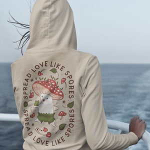 The back of a long-sleeved sand color hoodie. It has an art imprint of the colorful mushroom Amanita Muscaria in cartoon form and the words Spread Love Like Spores around the art.