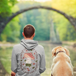 The back of a long-sleeved ash color hoodie. It has an art imprint of the colorful mushroom Amanita Muscaria in cartoon form and the words Spread Love Like Spores around the art. Photo also includes an incredibly cute lab dog.