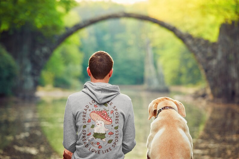 The back of a long-sleeved ash color hoodie. It has an art imprint of the colorful mushroom Amanita Muscaria in cartoon form and the words Spread Love Like Spores around the art. Photo also includes an incredibly cute lab dog.