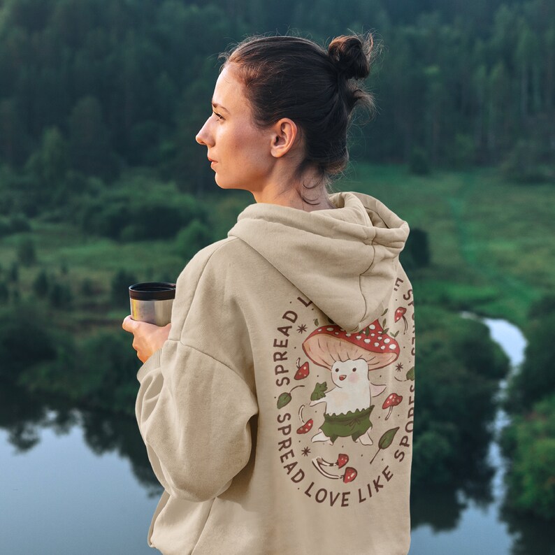 The back of a long-sleeved sand color hoodie. It has an art imprint of the colorful mushroom Amanita Muscaria in cartoon form and the words Spread Love Like Spores around the art.