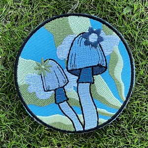 May include: A round embroidered patch with a blue background and two white mushrooms with blue caps. The mushrooms have green and white flowers on top. The background has a green and white swirl pattern.