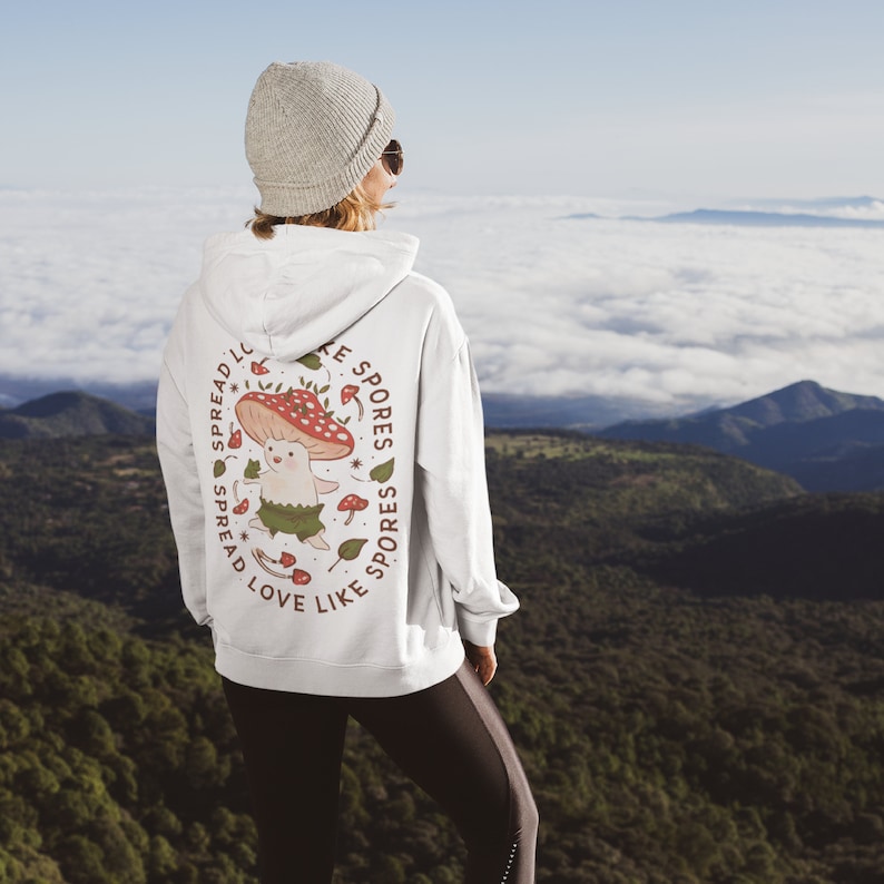 The back of a long-sleeved white color hoodie. It has an art imprint of the colorful mushroom Amanita Muscaria in cartoon form and the words Spread Love Like Spores around the art.