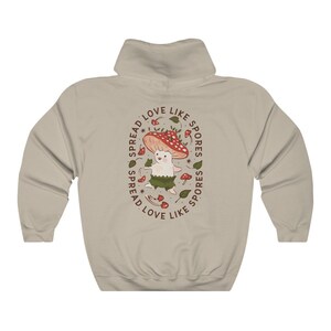 The back of a long-sleeved sand color hoodie. It has an art imprint of the colorful mushroom Amanita Muscaria in cartoon form and the words Spread Love Like Spores around the art.