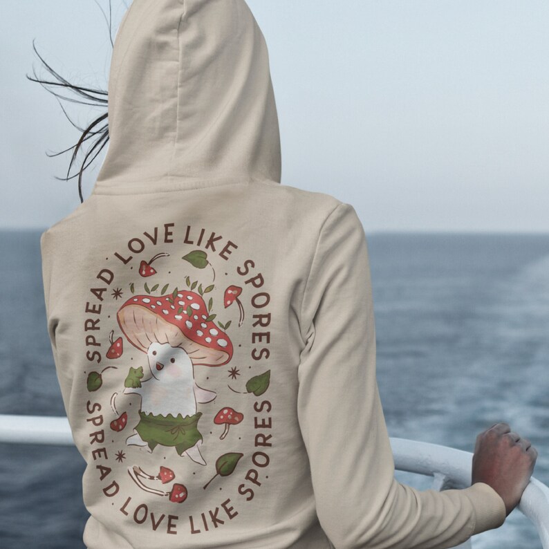 The back of a long-sleeved sand color hoodie. It has an art imprint of the colorful mushroom Amanita Muscaria in cartoon form and the words Spread Love Like Spores around the art.