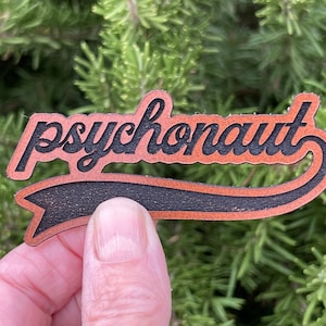 May include: A wooden cutout with the word "psychonaut" in a stylized font. The word is in a light brown color with a black outline, set against a black banner. The cutout is held against a backdrop of green foliage.