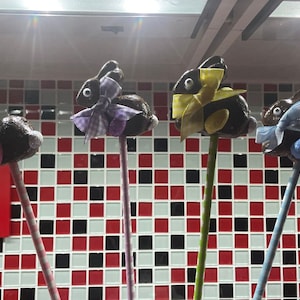 May include: Four chocolate bunny pops decorated with colorful bows and ribbons. The bunnies are on sticks and are arranged in a row on a tiled surface.