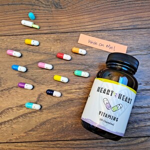 May include: A glass bottle of Heart 2 Heart vitamins with a label that says "Vitamins (60 Capsules)". The bottle is surrounded by colorful capsules with smiley faces on them. A sticky note says "Write on Me!"