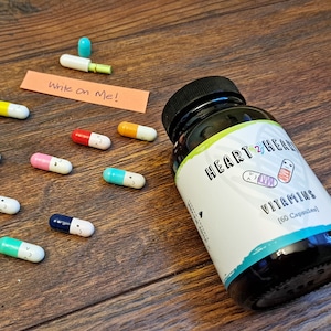 May include: A black bottle of "Heart 2 Heal Vitamins" with a white label and a blue border. Scattered around are colorful capsule-shaped vitamins with smiling faces. A small orange sticky note reads "Write on Me!"