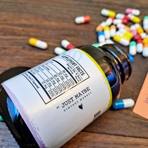 May include: A white bottle with a black label that says "Supplement Facts" and lists the amount per serving of various ingredients. The label also says "By Just Maybe Vintage Market". The bottle is lying on a wooden surface with a pile of colorful pills scattered around it.