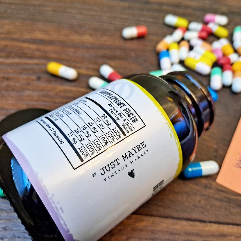 May include: A white bottle with a black label that says "Supplement Facts" and lists the amount per serving of various ingredients. The label also says "By Just Maybe Vintage Market". The bottle is lying on a wooden surface with a pile of colorful pills scattered around it.