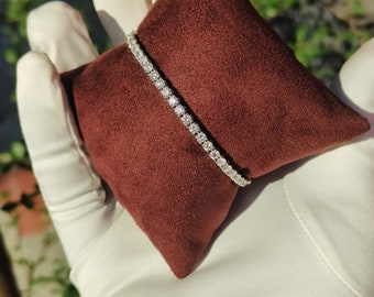 May include: A white gold tennis bracelet with a row of round brilliant diamonds set in a prong setting. The bracelet is displayed on a brown velvet cushion.