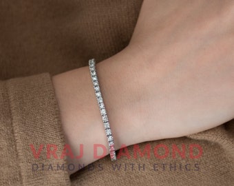May include: A tennis bracelet with numerous round-cut diamonds set in a row. The bracelet is silver-toned and is worn on a person's wrist. The background is a brown sweater. The text "VRAJ DIAMOND" and "DIAMONDS WITH ETHICS" is visible.