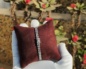 May include: A silver tennis bracelet with a row of clear cubic zirconia stones on a brown velvet cushion.