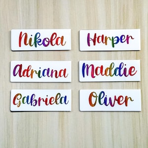 May include: Six white rectangular name tags with rainbow-coloured lettering. The names are Nikola, Harper, Adriana, Maddie, Gabriela, and Oliver. The names are written in a cursive font.