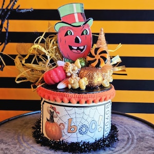 May include: A cylindrical Halloween decoration featuring a vintage-style pumpkin character in a green top hat, surrounded by straw, faux candy, and glittery accents. The base is wrapped in white paper with a spiderweb print, a black cat, and the word "boo!"