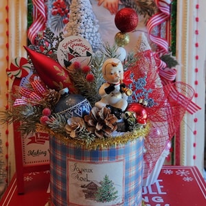 May include: A festive Christmas decoration featuring a blue plaid cylinder adorned with a vintage image and the words "Joyeux Noel." It is embellished with ornaments, pinecones, a small figurine, and a "Season's Greetings" tag. Red and gold accents complete the holiday theme.
