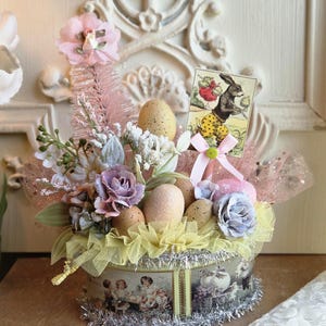May include: An Easter-themed decorative arrangement in a round container. The arrangement includes speckled eggs, artificial flowers in pastel colors, and a vintage-style card featuring a rabbit. The container is adorned with a vintage illustration and silver tinsel.