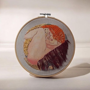 May include: Embroidered artwork in a round wooden hoop. The image depicts a nude figure with red hair, curled up in a fetal position. The embroidery uses various colors and textures to create depth and detail in the artwork.