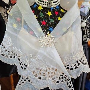 以下が含まれることがあります： 複雑な花柄の刺繍とスカラップ状のカットワークの裾が特徴の、透け感のある白いオーガンジーのショール。ショールの中央は小さな白い真珠のクラスターで留められており、多連の真珠のネックレスを身に着けたマネキンに掛けられています。