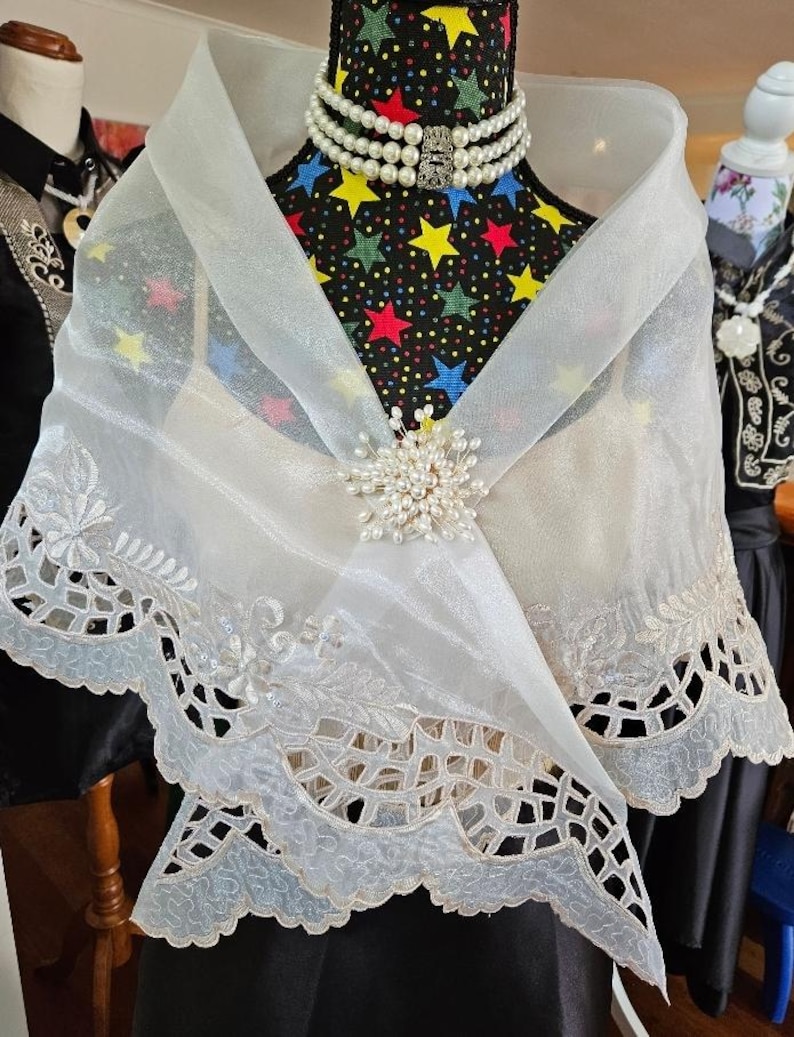 以下が含まれることがあります： 複雑な花柄の刺繍とスカラップ状のカットワークの裾が特徴の、透け感のある白いオーガンジーのショール。ショールの中央は小さな白い真珠のクラスターで留められており、多連の真珠のネックレスを身に着けたマネキンに掛けられています。