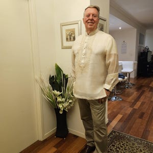 May include: A cream-colored, long-sleeved barong tagalog shirt featuring a mandarin collar, a button-down front placket, and intricate tonal embroidery on the chest, paired with light brown trousers and dark brown leather dress shoes.