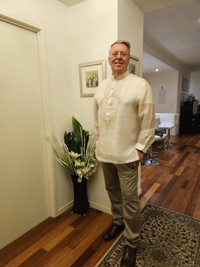 May include: A cream-colored, long-sleeved barong tagalog shirt featuring a mandarin collar, a button-down front placket, and intricate tonal embroidery on the chest, paired with light brown trousers and dark brown leather dress shoes.