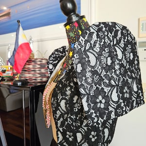 May include: A black lace bolero jacket featuring prominent, structured butterfly sleeves and a floral pattern. The garment is displayed on a mannequin, showcasing the intricate lace overlay and the distinct, wide-shouldered silhouette of the sleeves.