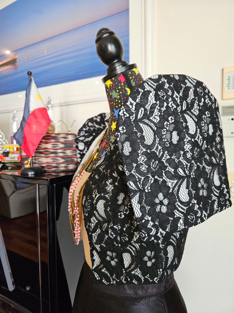 May include: A black lace bolero jacket featuring prominent, structured butterfly sleeves and a floral pattern. The garment is displayed on a mannequin, showcasing the intricate lace overlay and the distinct, wide-shouldered silhouette of the sleeves.