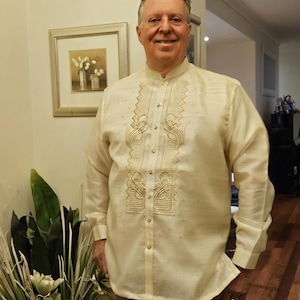 May include: A long-sleeved, cream-colored Barong Tagalog shirt featuring a mandarin collar, a button-down front, and intricate, symmetrical embroidery patterns along the chest panel. The fabric has a subtle, semi-sheer, and textured appearance.