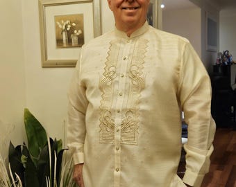 May include: A long-sleeved, cream-colored Barong Tagalog shirt featuring a mandarin collar, a button-down front, and intricate, symmetrical embroidery patterns along the chest panel. The fabric has a subtle, semi-sheer, and textured appearance.