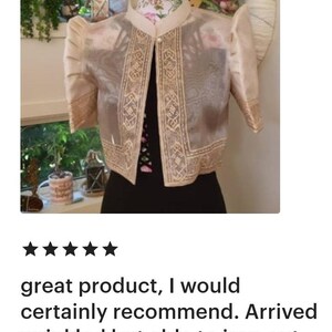 May include: A beige bolero jacket featuring sheer fabric, structured puff sleeves, and a mandarin collar. The garment is adorned with intricate gold-toned embroidery along the front edges and sleeve cuffs, secured by a single pearl-like button closure.