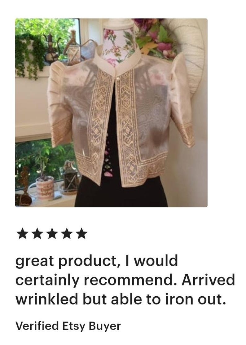 May include: A beige bolero jacket featuring sheer fabric, structured puff sleeves, and a mandarin collar. The garment is adorned with intricate gold-toned embroidery along the front edges and sleeve cuffs, secured by a single pearl-like button closure.