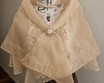 May include: A sheer beige organza shawl featuring delicate floral embroidery along the scalloped edges, secured at the centre with a round gold-tone brooch adorned with small white pearls and clear crystals, draped over a mannequin with a pearl necklace.