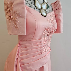 May include: A light pink formal gown with a sweetheart neckline and pleated bodice. The dress features embellished floral embroidery and pearl accents. A statement necklace with large, white, teardrop-shaped pendants adorns the mannequin.