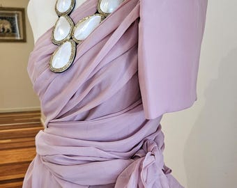 May include: A lavender one-shoulder dress featuring draped fabric, a large structured sleeve, and a gathered waist with a side tie. A statement necklace with five large, white, teardrop-shaped stones set in gold-toned trim rests on the neckline.
