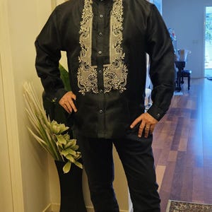 May include: A black long-sleeved button-down shirt featuring a mandarin collar and intricate cream-colored embroidery along the front placket. The shirt is paired with matching black trousers and black dress shoes, creating a formal ensemble.