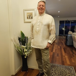 May include: A cream-colored long-sleeved barong tagalog shirt featuring a mandarin collar, a button-down front placket, and intricate tonal embroidery on the chest, paired with light khaki trousers and dark brown leather dress shoes.