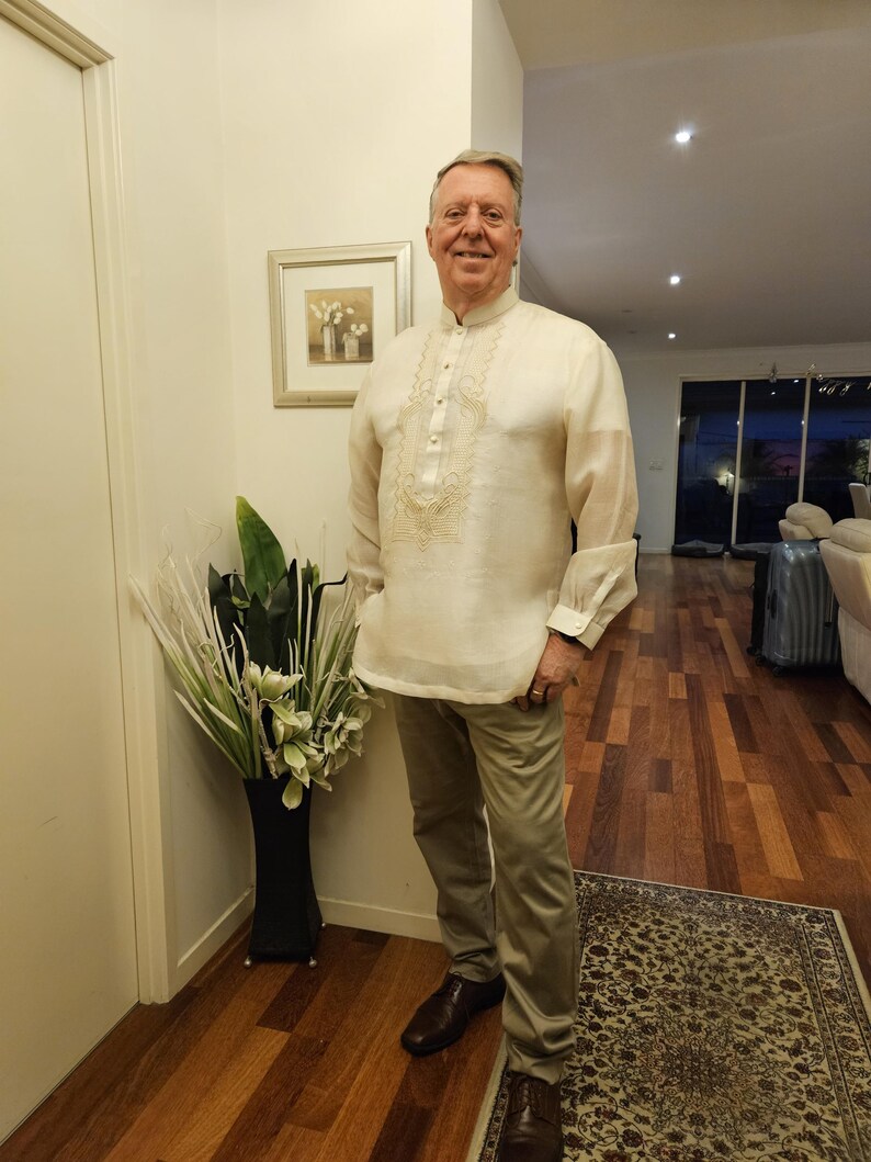 May include: A cream-colored long-sleeved barong tagalog shirt featuring a mandarin collar, a button-down front placket, and intricate tonal embroidery on the chest, paired with light khaki trousers and dark brown leather dress shoes.