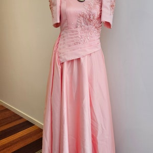 May include: A light pink formal gown with short sleeves and a floor-length skirt. The dress features intricate beadwork and embroidery on the bodice and sleeves. A statement necklace with large, white, shell-shaped pendants adorns the neckline.
