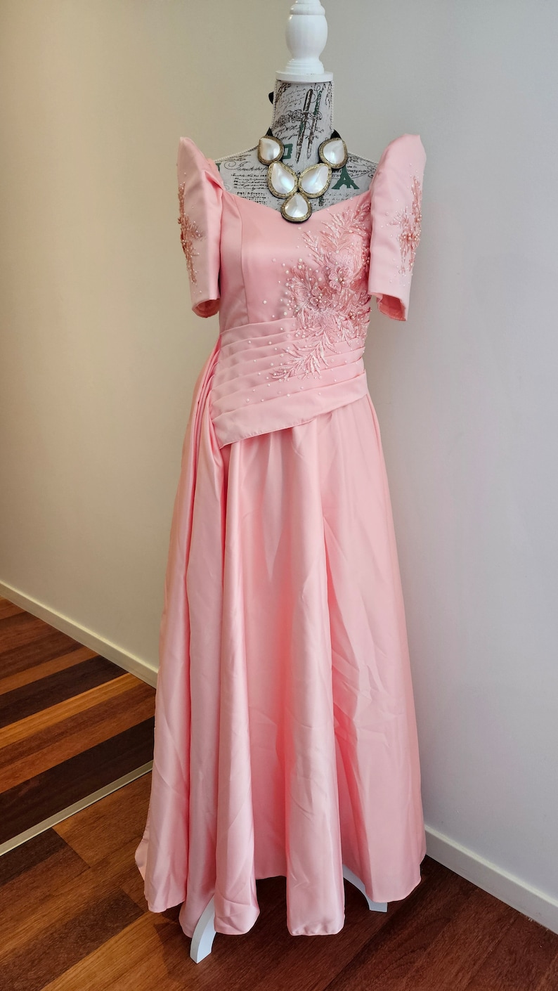 May include: A light pink formal gown with short sleeves and a floor-length skirt. The dress features intricate beadwork and embroidery on the bodice and sleeves. A statement necklace with large, white, shell-shaped pendants adorns the neckline.