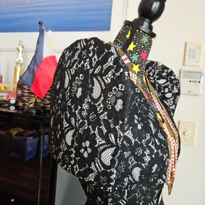 May include: A black lace bolero jacket with prominent puffed sleeves and a floral pattern, displayed on a mannequin. The garment features a structured shoulder design and is layered over a solid black skirt, accented by a long beaded necklace.