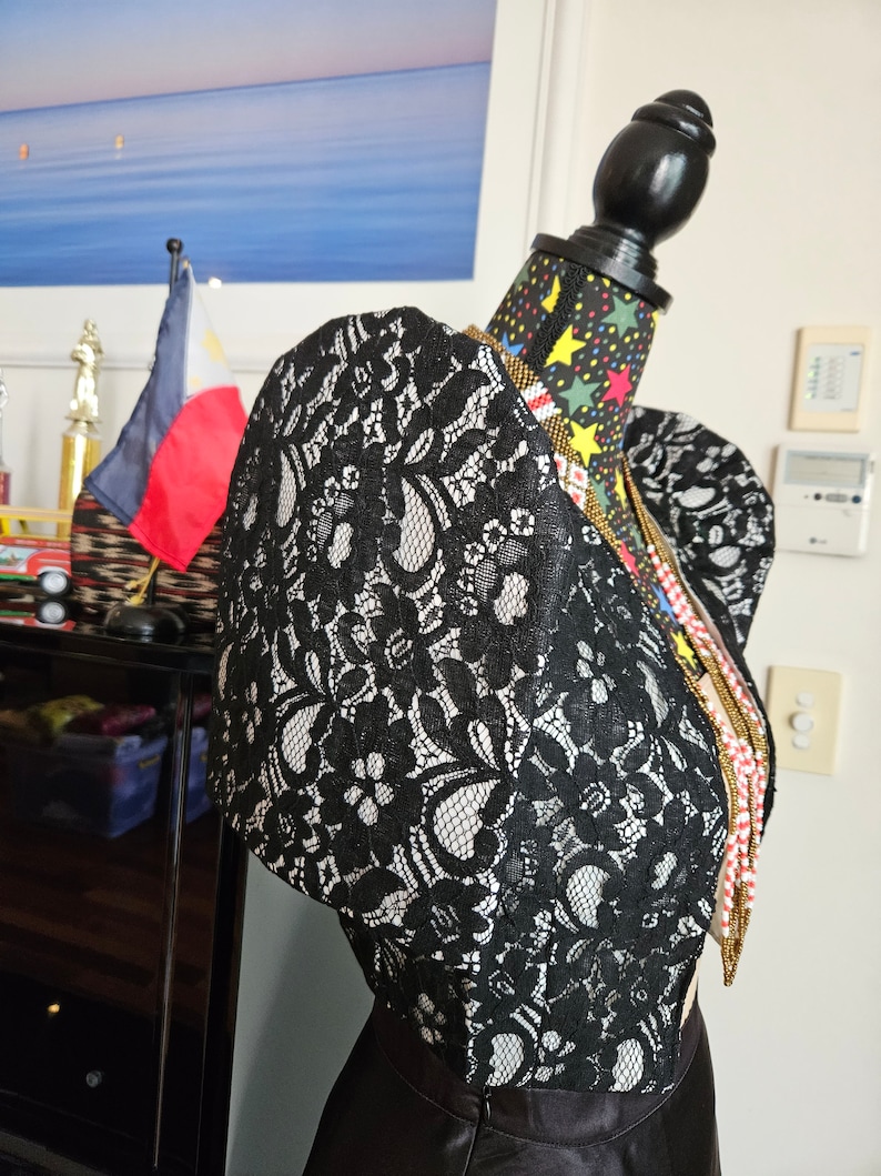 May include: A black lace bolero jacket with prominent puffed sleeves and a floral pattern, displayed on a mannequin. The garment features a structured shoulder design and is layered over a solid black skirt, accented by a long beaded necklace.