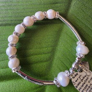 Peut inclure: Un bracelet de perles avec des d&eacute;tails argent&eacute;s et un charm en forme de c&oelig;ur. Le bracelet pr&eacute;sente de grosses perles blanc cass&eacute; s&eacute;par&eacute;es par des entretoises argent&eacute;es avec des cristaux clairs. Un charm en forme de c&oelig;ur avec du texte grav&eacute; est suspendu.