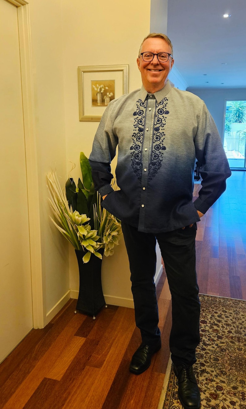 May include: A long-sleeved button-down shirt featuring an ombre effect that transitions from light blue at the shoulders to dark blue at the hem. The shirt includes a vertical panel of dark blue floral embroidery along the center front placket.